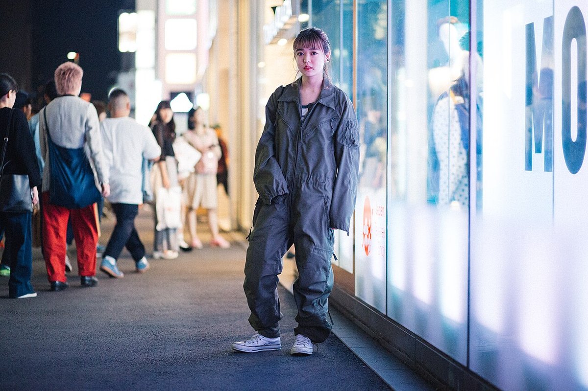 Woman standing on a city street at night wearing a charcoal grey utility jumpsuit with a belted waist and white trainers