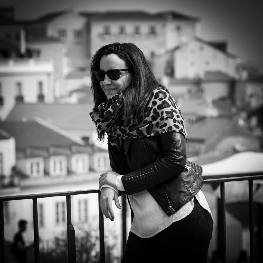 Black-and-white photo of a woman wearing sunglasses, a leopard-print scarf, a black leather biker jacket and dark jeans, leaning on a railing in Lisbon.