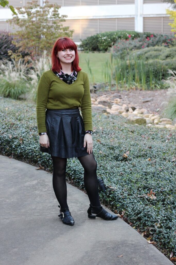 Red-haired woman outdoors wearing an olive green v-neck sweater over a floral shirt with a black pleated skirt and tights