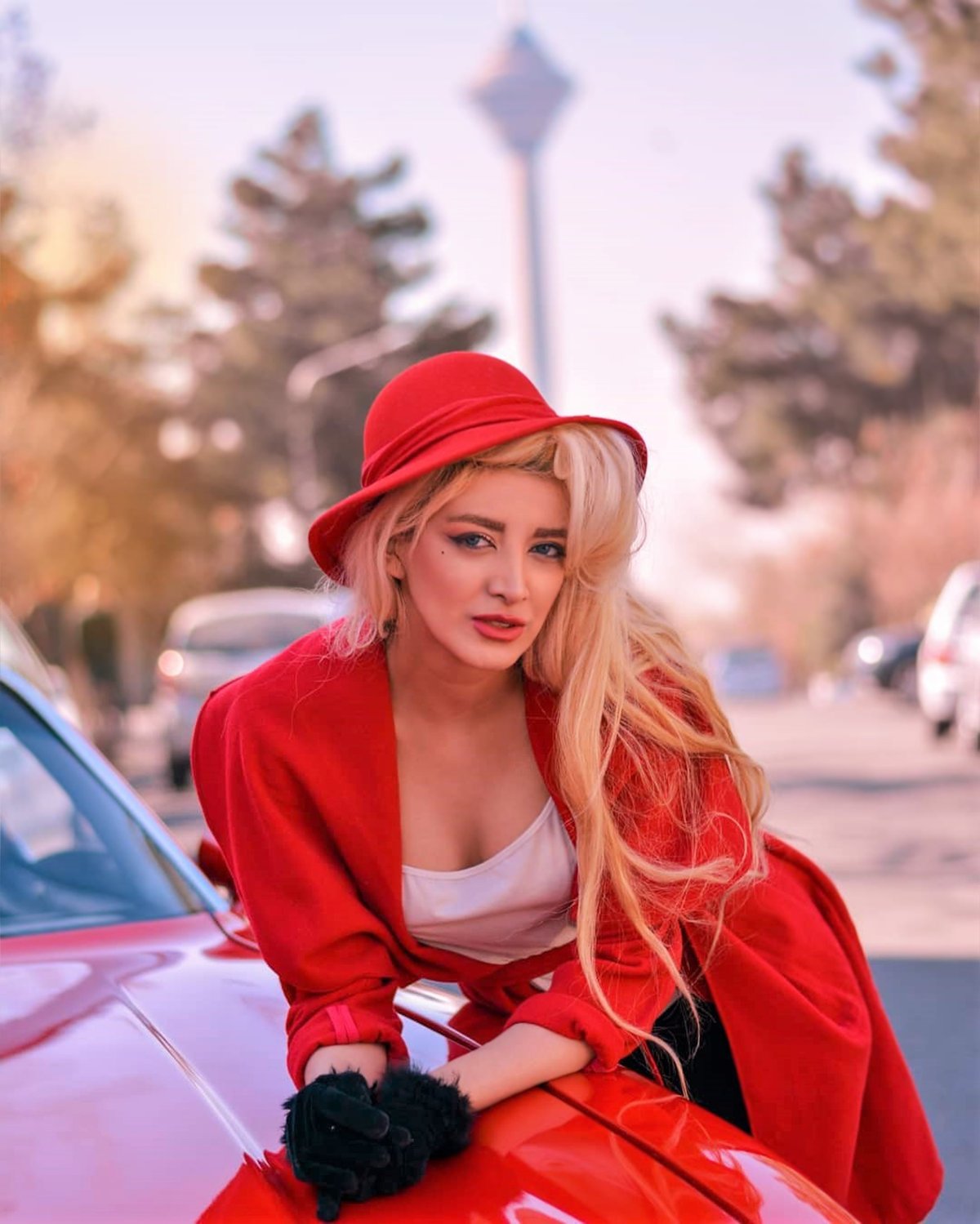 Woman in a red coat and matching red cloche hat with black fur gloves, leaning on a red car