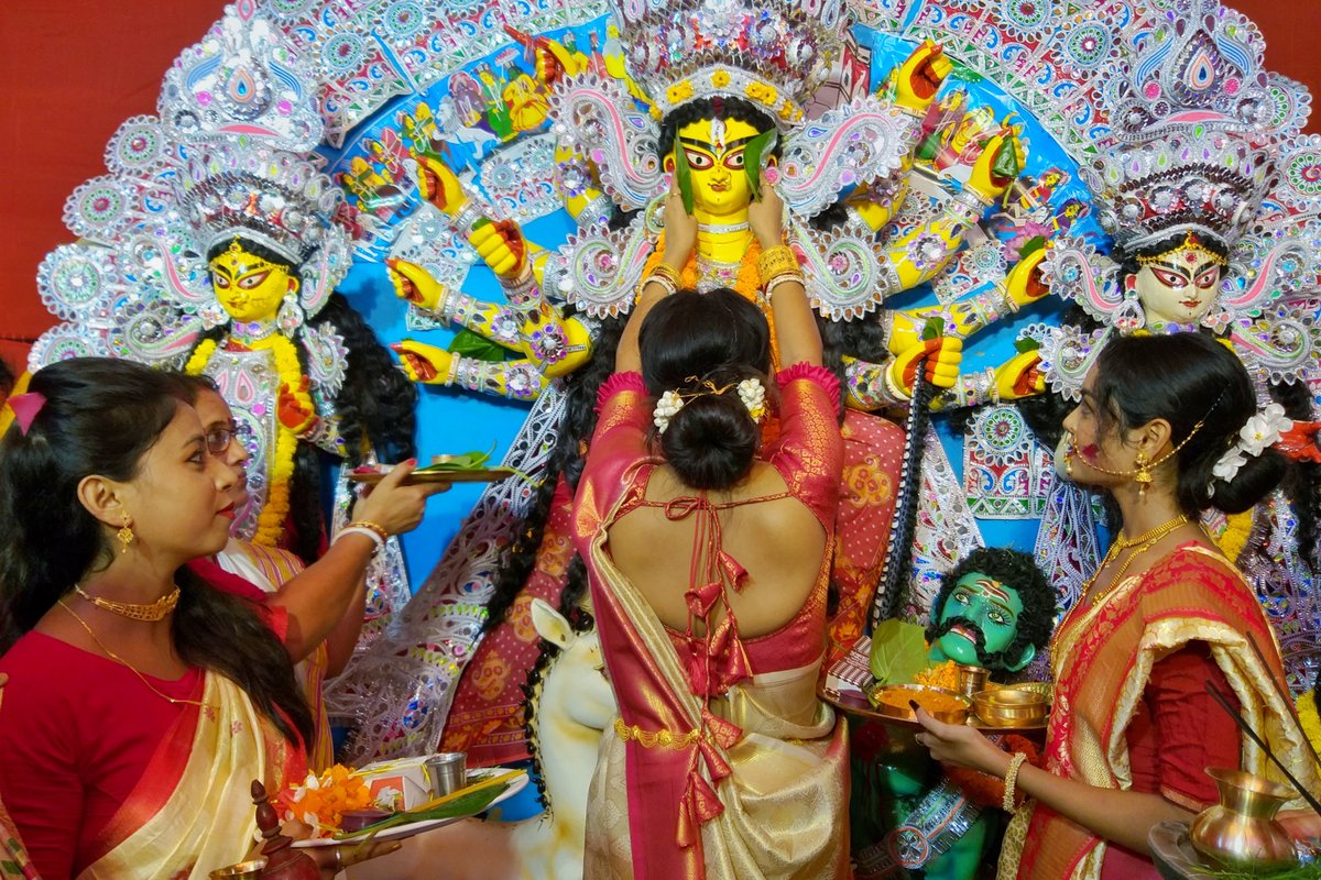 Three women in red blouses and cream and red sarees holding puja thalis and performing boron rituals in front of a large Durga idol