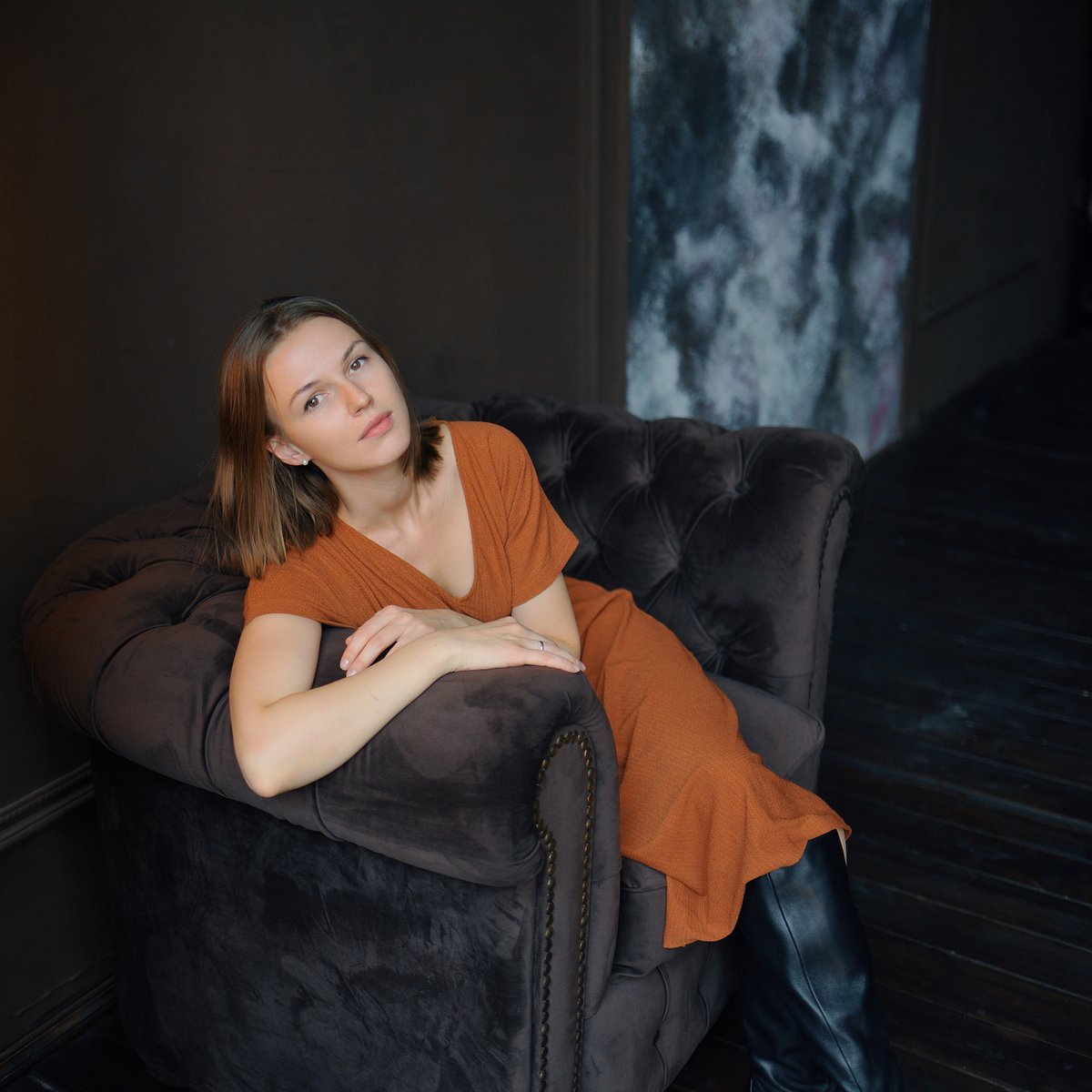 Woman reclining on a dark armchair wearing a rust-colored ribbed knit dress and black knee-high boots