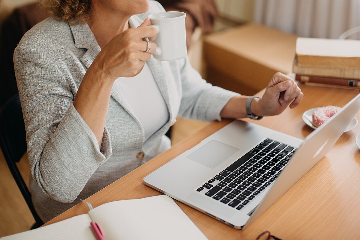 Woman in a gray blazer drinking coffee while working on a laptop at a wooden desk