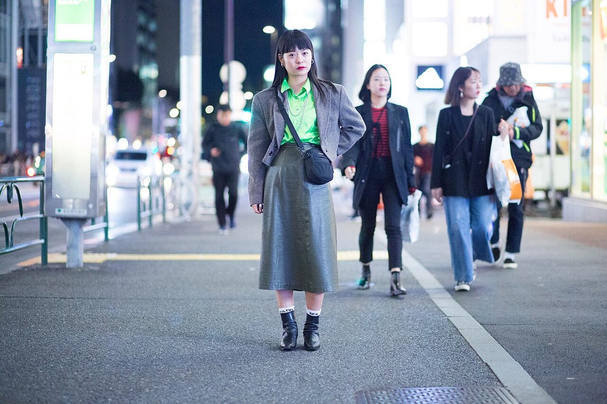 Woman standing on a night street wearing a cropped grey jacket over a bright green shirt with a long grey-green flowing skirt and ankle boots