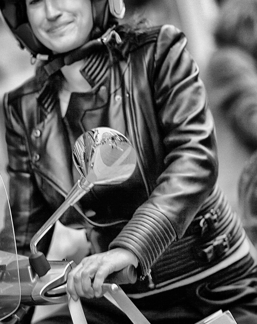 Woman in a leather jacket and helmet riding a Vespa scooter.