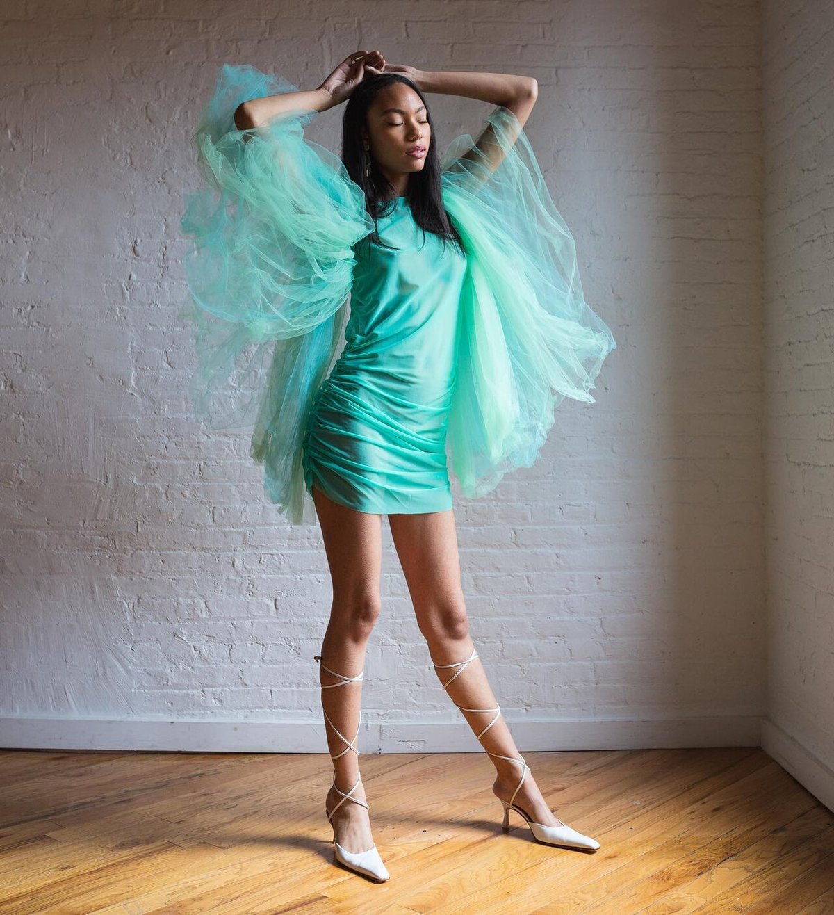 Woman standing indoors wearing a mint-teal ruched mini dress with voluminous sheer tulle sleeves and strappy heels