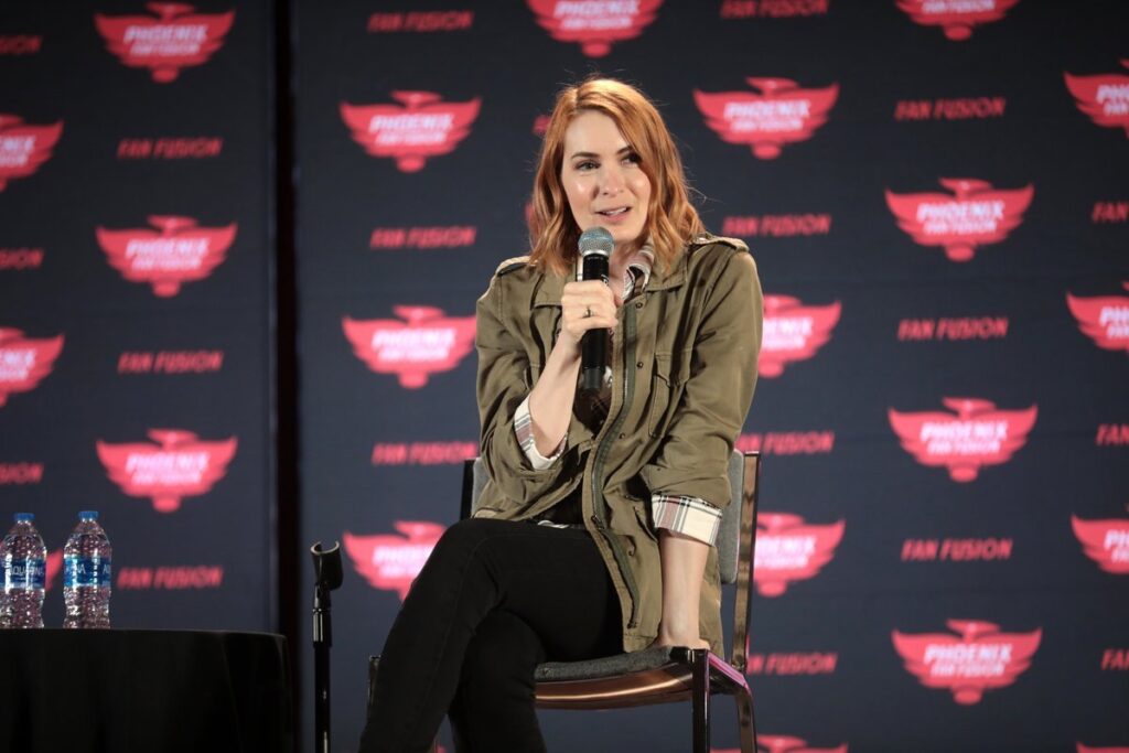 Felicia Day seated at a convention panel wearing an olive utility jacket over a plaid shirt, holding a microphone