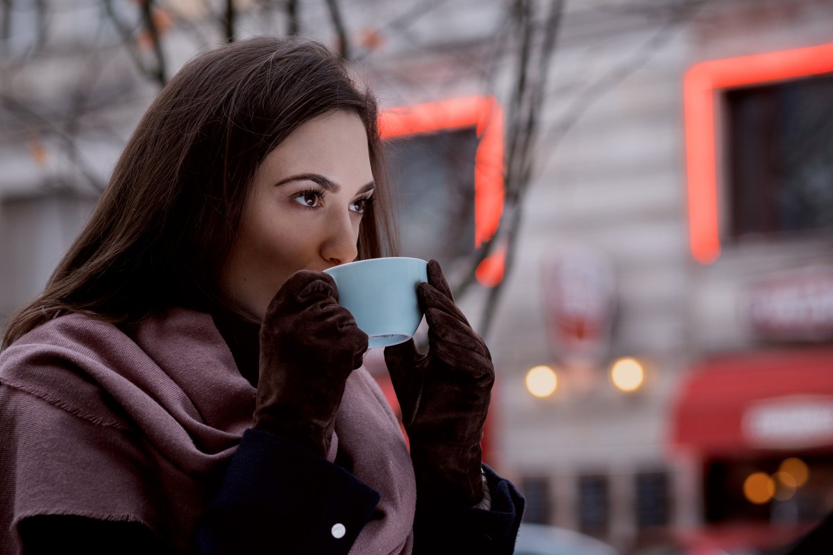 Woman in a mauve scarf drinking coffee outdoors wearing brown suede gloves