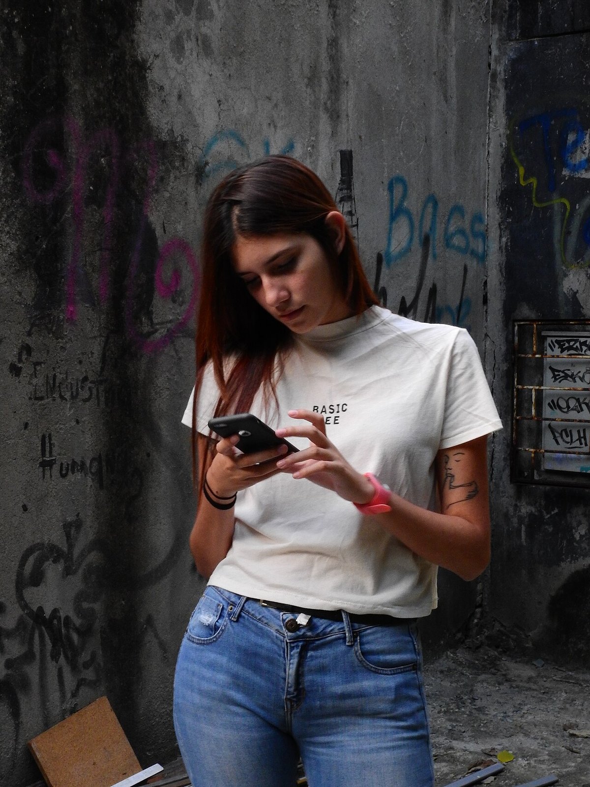 Woman on a city street looking at her phone, wearing a white printed T-shirt tucked into mid-blue jeans with a studded belt
