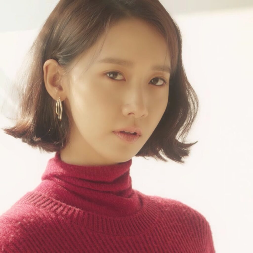 Woman with a short dark bob and gold hoop earrings in a bright editorial portrait, wearing a rich wine-burgundy ribbed turtleneck