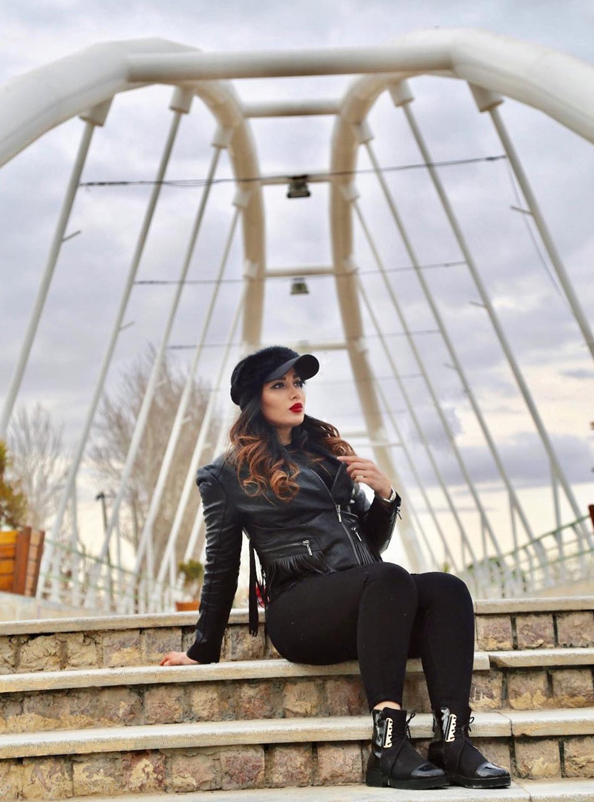 Woman seated on bridge steps in a black quilted leather jacket, black leggings, patent ankle boots and a black faux-fur cap