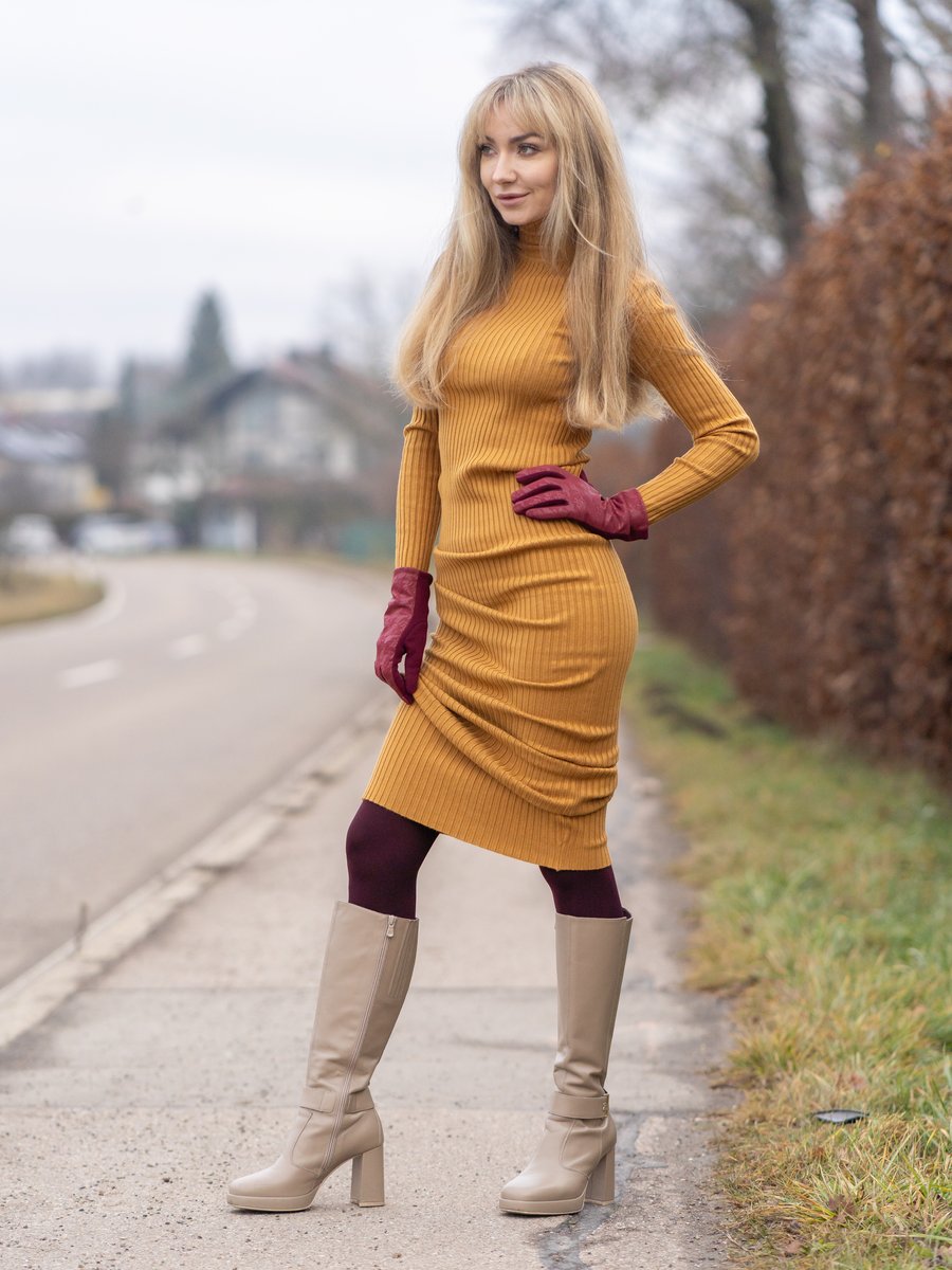 Blonde woman standing on a country path wearing a mustard-amber ribbed knit turtleneck midi dress with deep-burgundy leather gloves, opaque burgundy tights, and beige knee-high boots