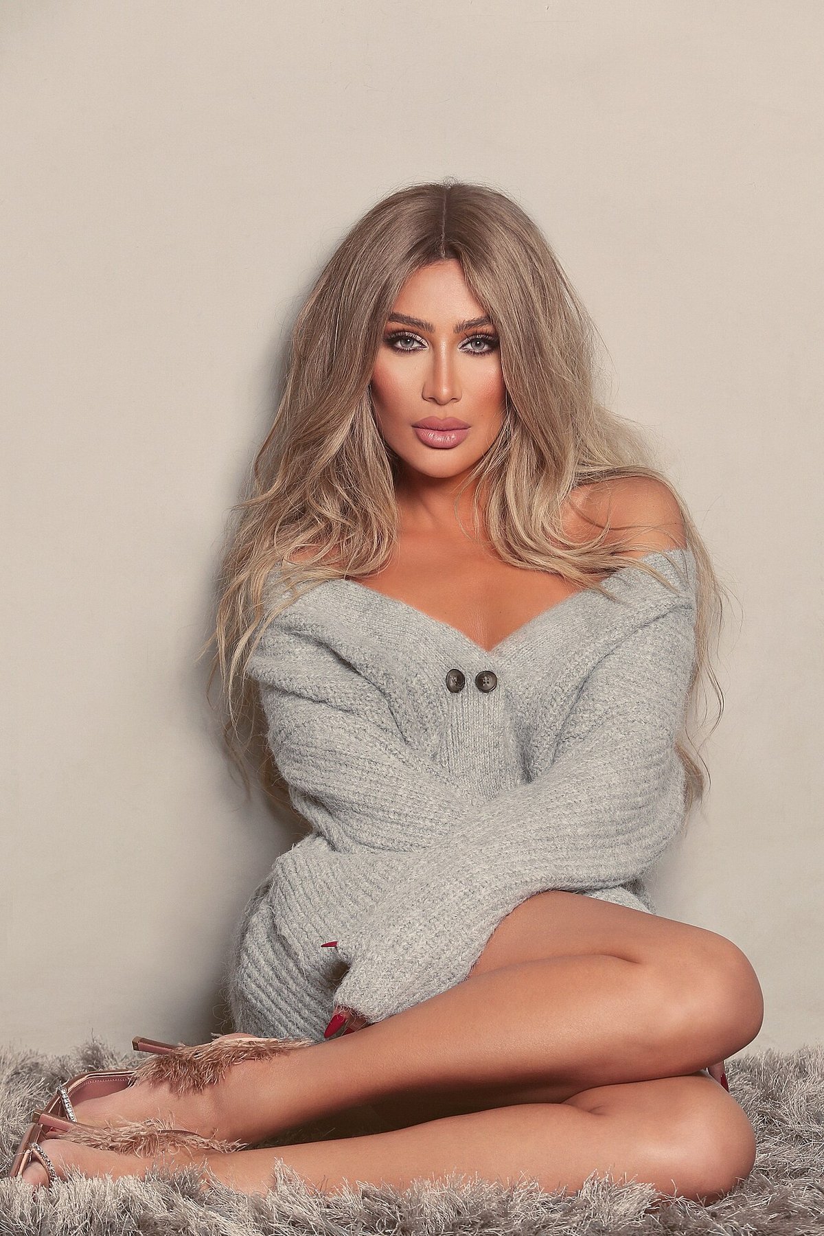 Portrait of a woman with long wavy hair wearing an off-shoulder light grey knitted cardigan dress with two dark buttons, seated cross-legged