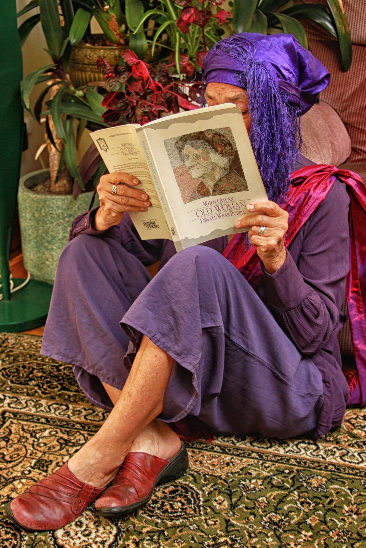 Older woman sitting indoors wearing a head-to-toe purple linen outfit with a purple headwrap and red shoes.