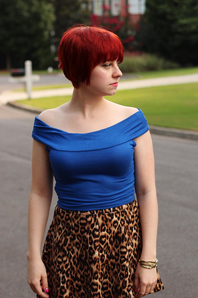 A woman with short red hair wearing a blue off-the-shoulder top and a knee-length flared leopard print skirt outdoors.