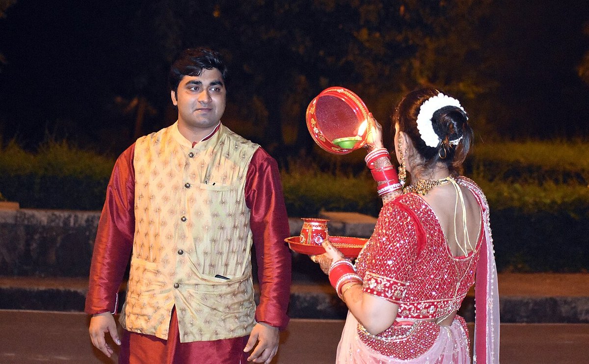 Back view of a woman in a red embroidered blouse and light saree with a white gajra in her bun, holding a decorated puja thali and sieve