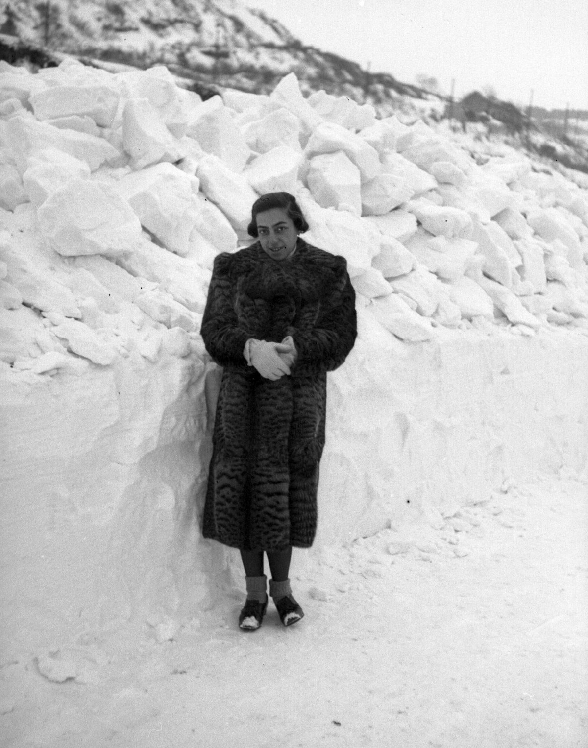 Vintage black-and-white photograph of a woman in a long fur coat and light gloves standing beside a tall wall of packed snow.