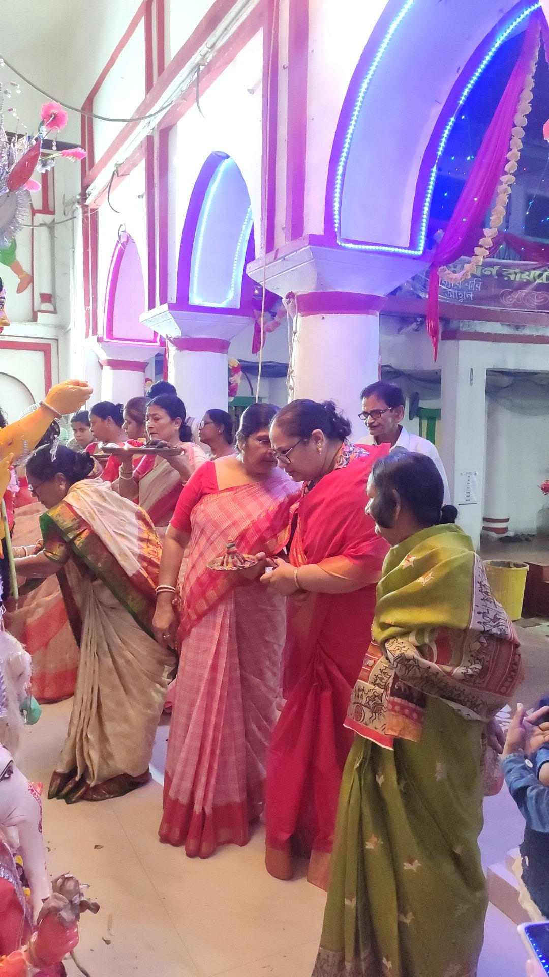 Women in red, pink and green sarees holding small boron thalis inside a decorated pandal during the Bijoya Dashami farewell ritual