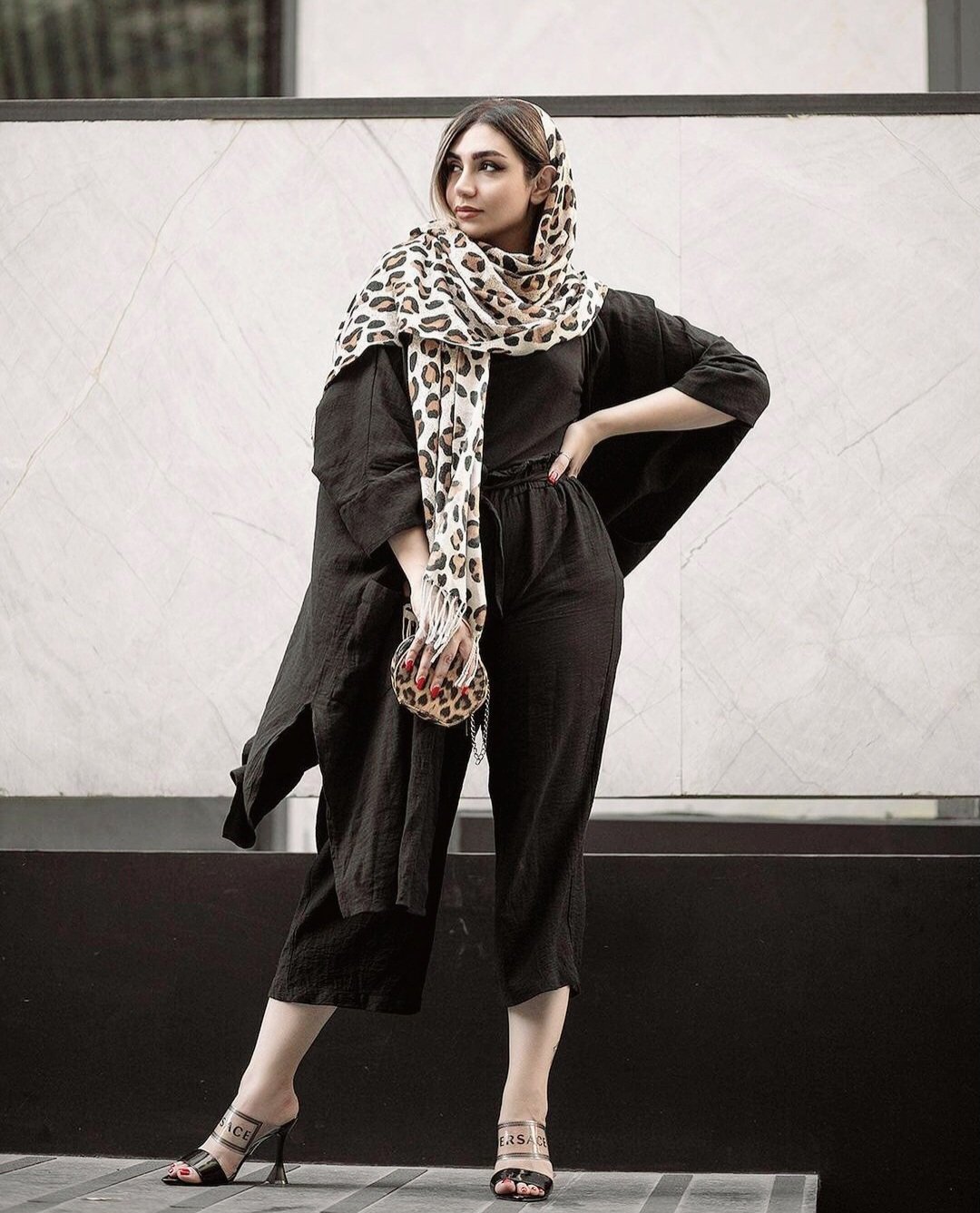 Woman in a black manteau and cropped black trousers with a leopard-print headscarf, leopard mini bag and heeled mules