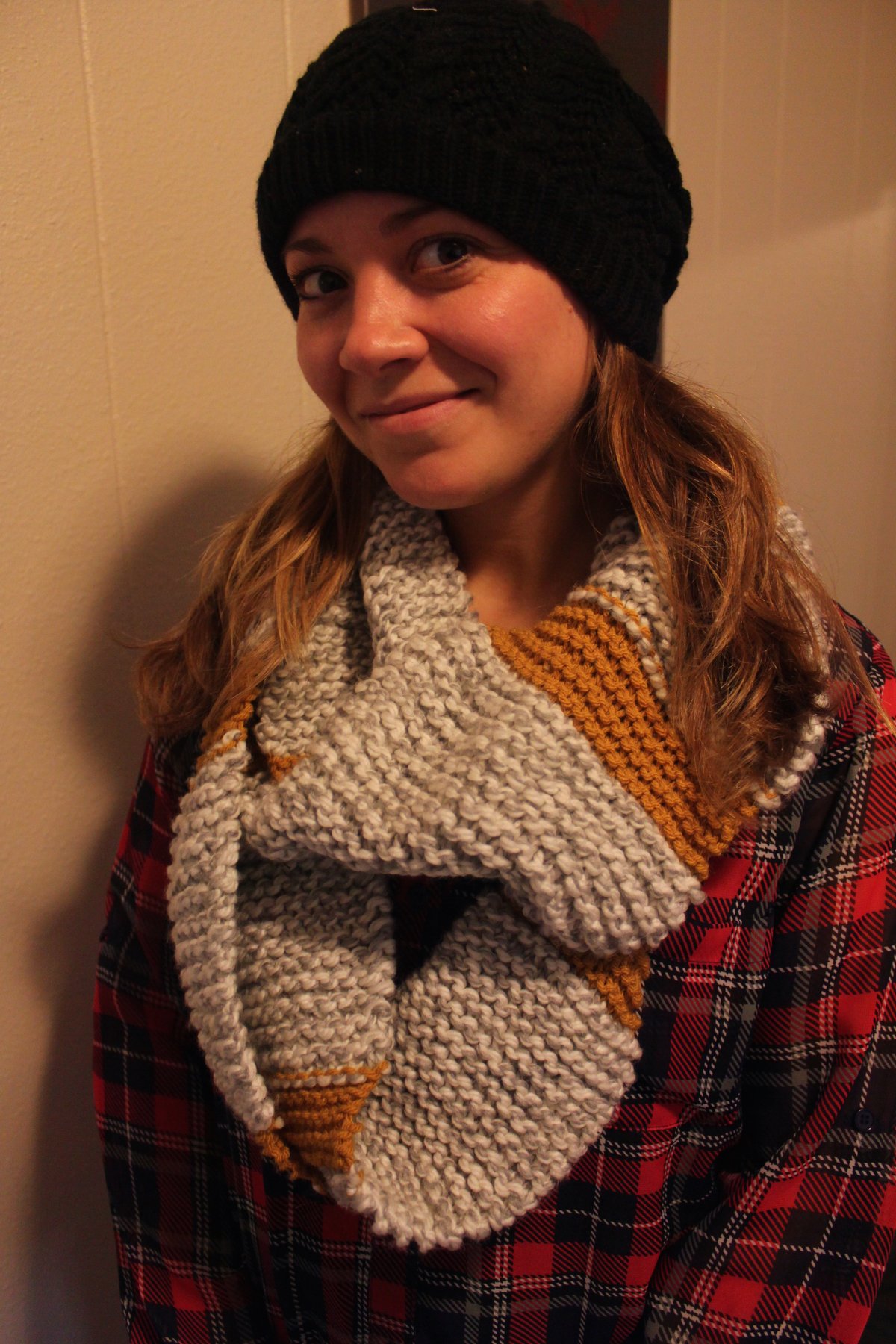 Woman indoors wearing a black knit beanie and a chunky grey and mustard striped knit infinity scarf over a red plaid shirt.