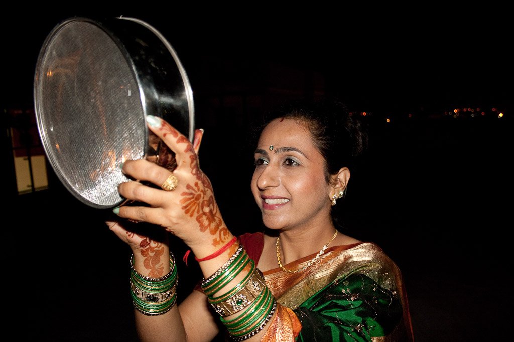 Woman at night holding a sieve for the moonrise ritual, wearing a green and gold saree with stacks of green and red bangles and henna on her hands