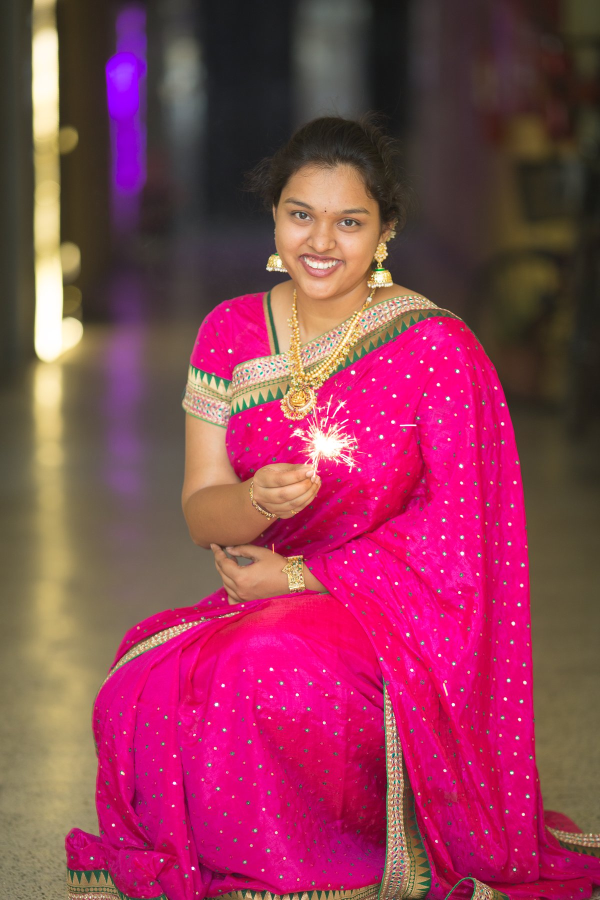 Woman in a magenta-pink embroidered saree with gold border and gold jewelry holding a lit sparkler
