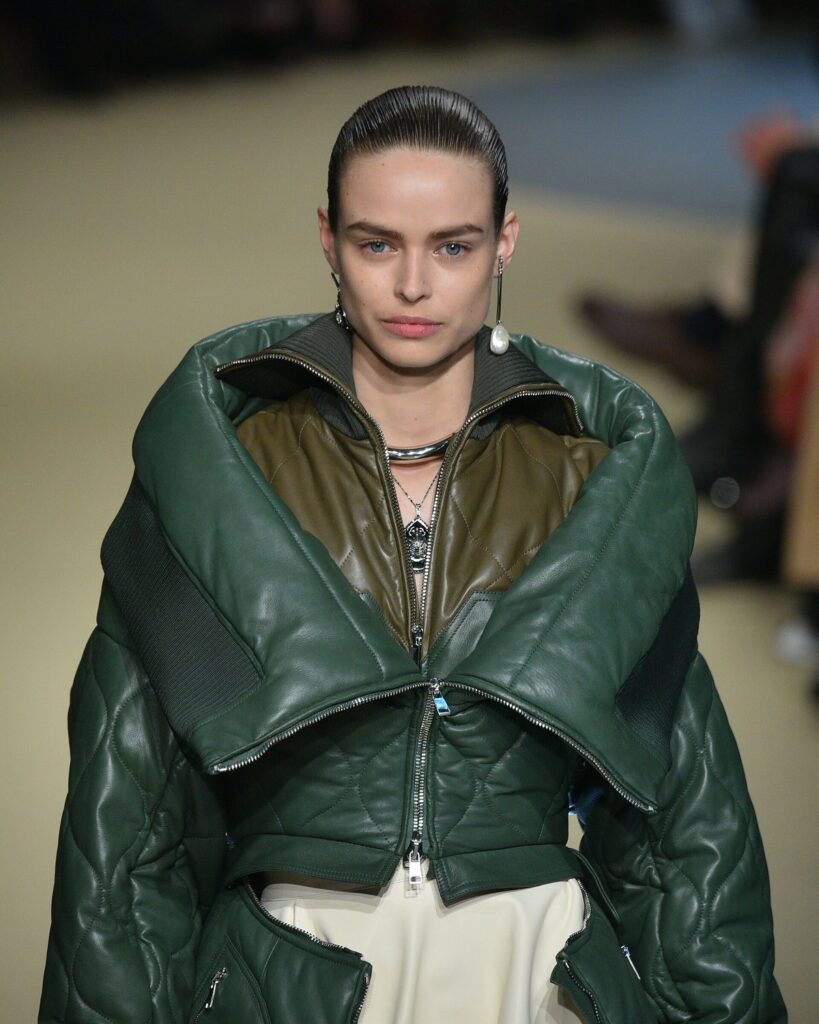 Runway model wearing a dark green quilted leather jacket with oversized lapels over a bronze quilted vest