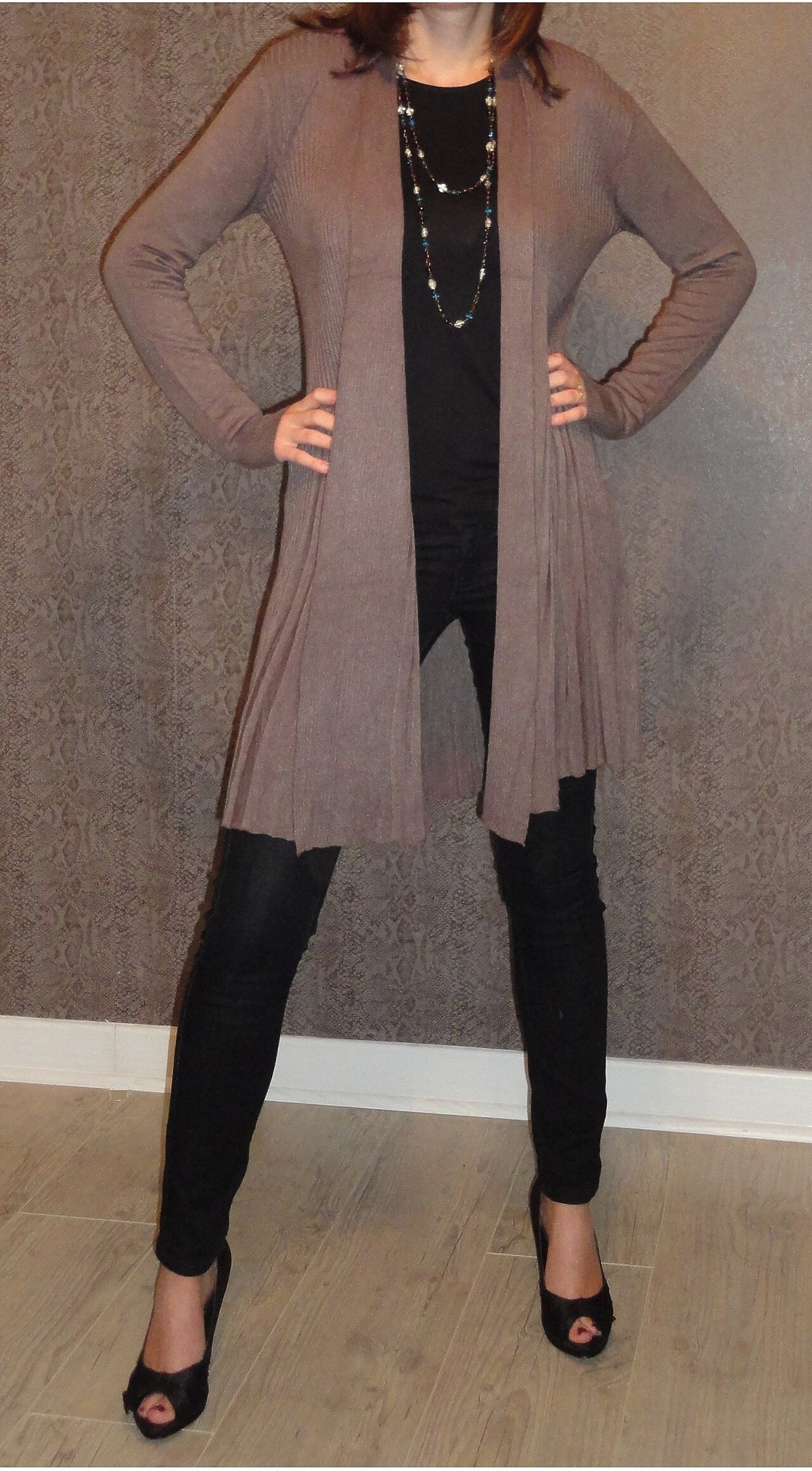 Woman standing indoors wearing a long taupe-grey pleated cardigan over a black top and black skinny trousers with layered necklaces and black heels