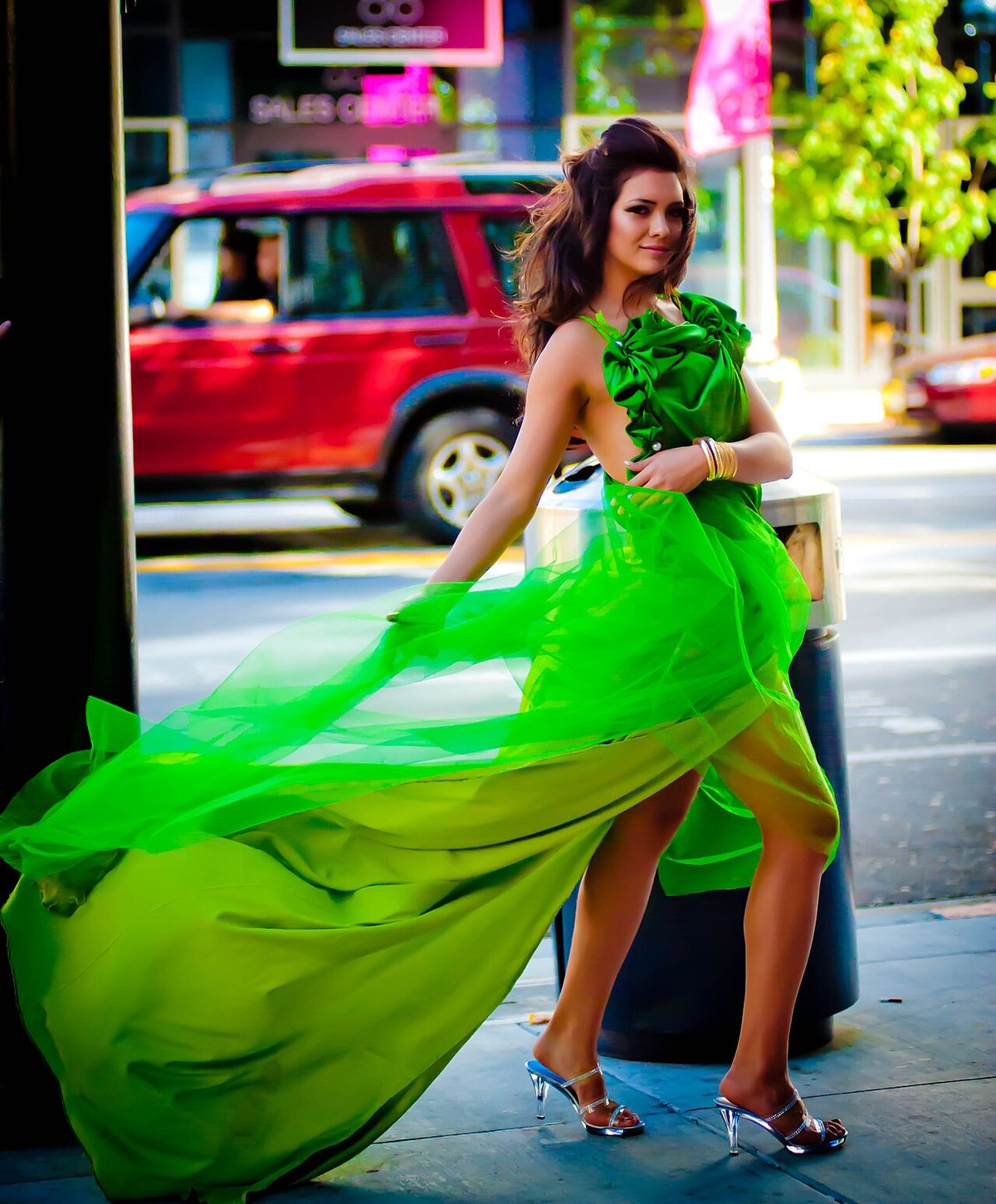 Woman on a city street holding out the flowing skirt of a vivid green gown with a ruffled bodice, the fabric caught mid-swirl
