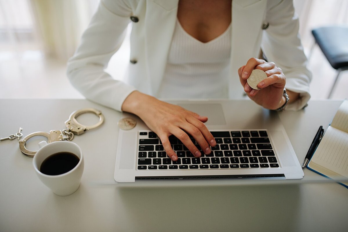 Woman in a white blazer typing on a laptop at a desk with a cup of coffee and handcuffs resting nearby