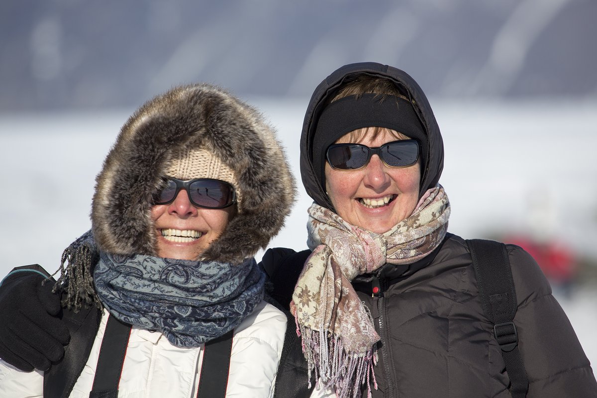 Two smiling women standing in snow wearing down jackets with fur-trimmed hoods, knit hats, sunglasses and scarves.
