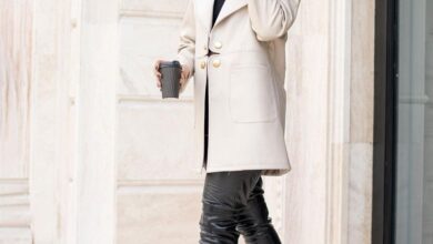 Woman in an ivory double-breasted coat with gold buttons, black leather leggings, black over-the-knee stiletto boots and a black fedora, holding a coffee cup