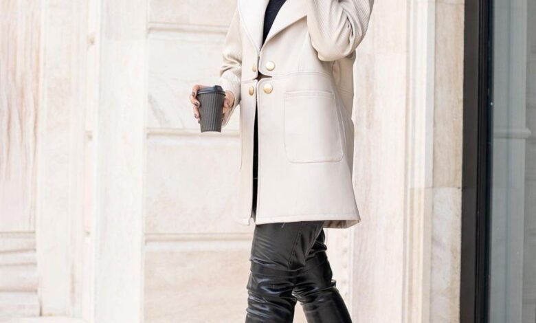 Woman in an ivory double-breasted coat with gold buttons, black leather leggings, black over-the-knee stiletto boots and a black fedora, holding a coffee cup