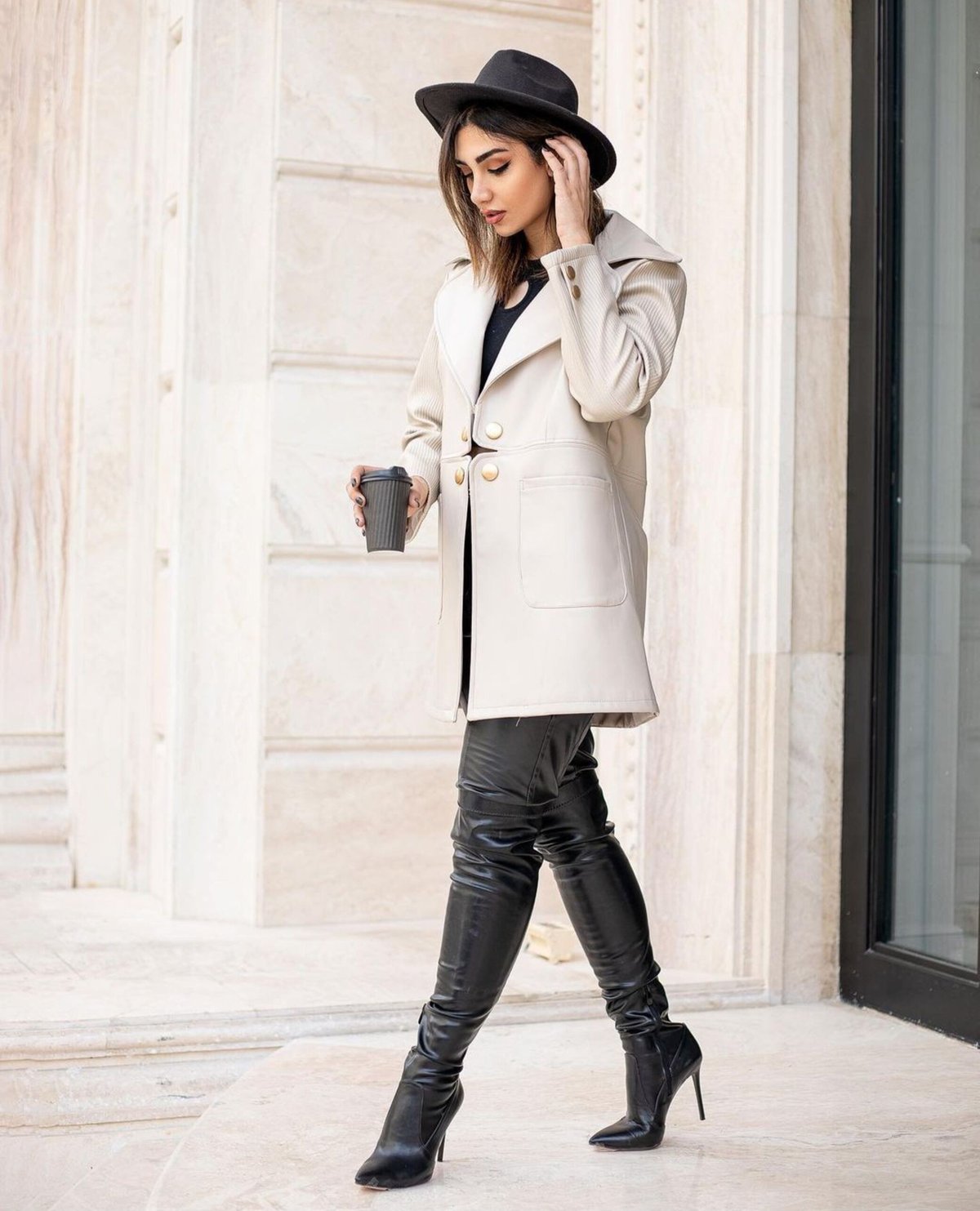 Woman in an ivory double-breasted coat with gold buttons, black leather leggings, black over-the-knee stiletto boots and a black fedora, holding a coffee cup