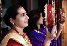 Two women holding decorated red sieves, the woman in front wearing a red saree with a cream and gold dupatta, a gold necklace and henna on her hands