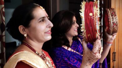 Two women holding decorated red sieves, the woman in front wearing a red saree with a cream and gold dupatta, a gold necklace and henna on her hands