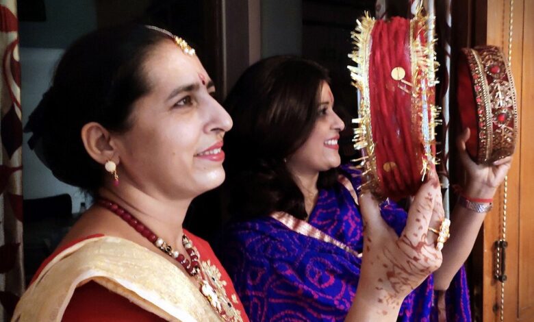 Two women holding decorated red sieves, the woman in front wearing a red saree with a cream and gold dupatta, a gold necklace and henna on her hands