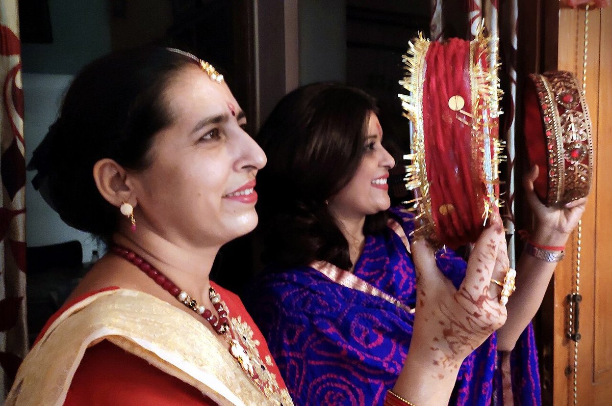 Two women holding decorated red sieves, the woman in front wearing a red saree with a cream and gold dupatta, a gold necklace and henna on her hands