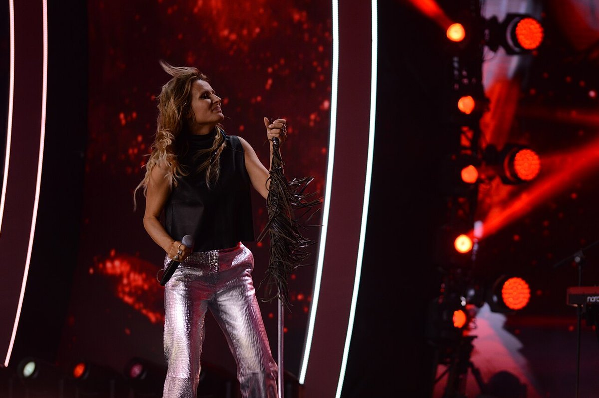 Woman performing on a stage in silver metallic trousers and a black sleeveless top, holding a microphone with stage lights behind her