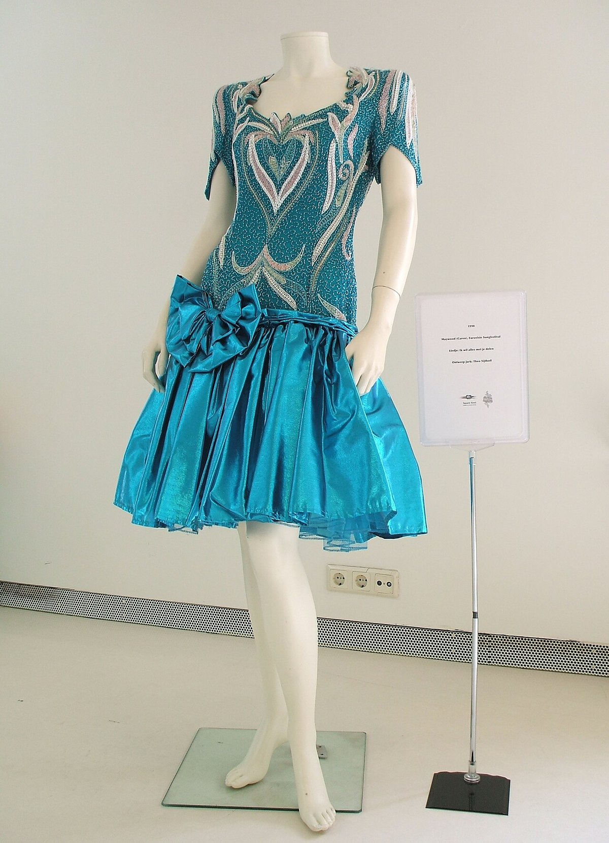 Teal dress with silver beaded embellishment, a sweetheart neckline and a large bow at the waist, displayed on a white mannequin in a museum
