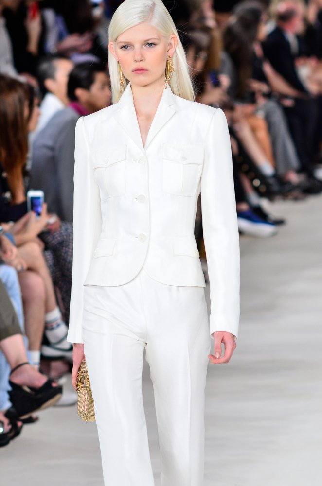 Runway model in an all-white pantsuit with patch-pocket blazer and slim white trousers, carrying a gold clutch