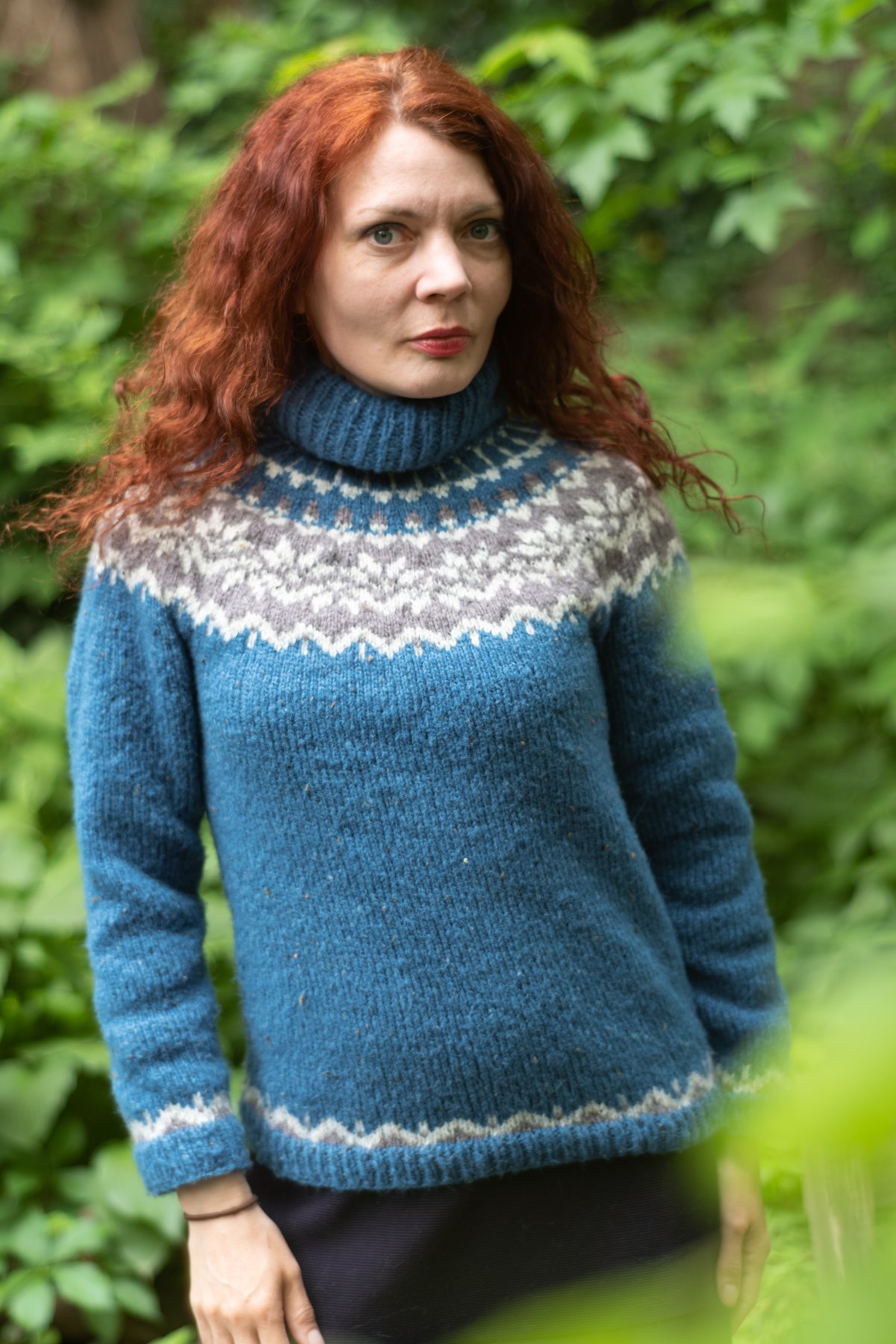 Woman outdoors wearing a blue Icelandic yoke-pattern knit sweater with a ribbed turtleneck collar.