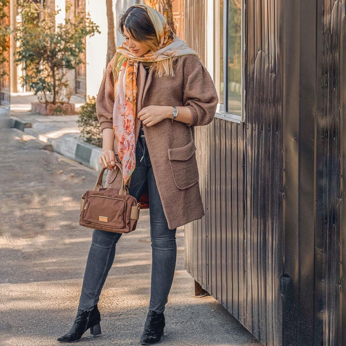 Woman on a street in a brown wool coat, dark jeans and black ankle boots, with a floral orange headscarf and brown handbag