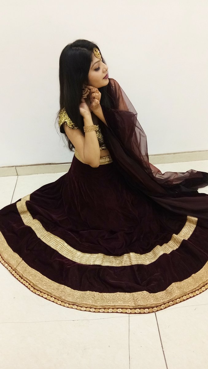 Young Indian woman seated on a tiled floor wearing a dark wine-burgundy velvet lehenga with wide gold embroidered borders, a gold sequined choli, and a sheer dark maroon dupatta over her head, with gold bangles and jhumka earrings
