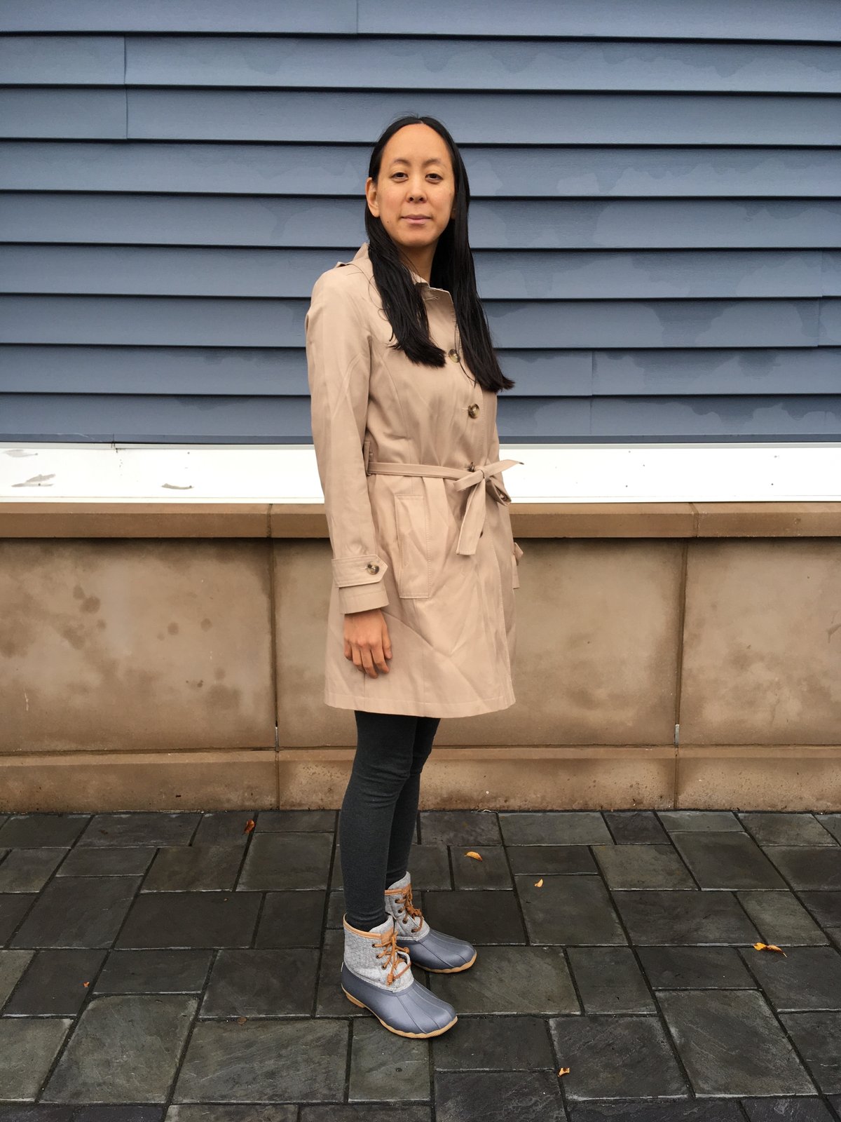 Woman with long dark hair outdoors wearing a beige belted trench coat with buttoned cuffs, over black leggings and lace-up duck boots