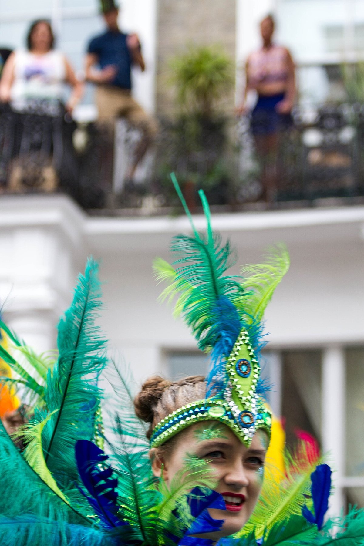 Close view of a carnival performer wearing an elaborate peacock feather headdress in teal, green and blue, with matching green and blue face paint