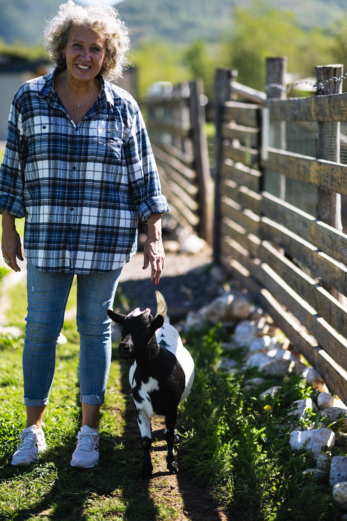 Woman walking along a wooden fence with a goat, wearing a blue plaid flannel shirt and blue jeans