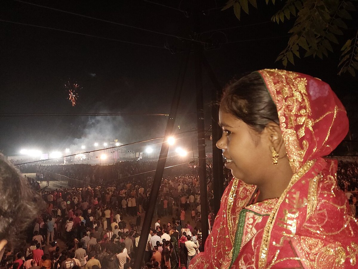 Profile of a young woman wearing a red saree with heavy gold embroidery and a matching dupatta over her head at a festive night gathering