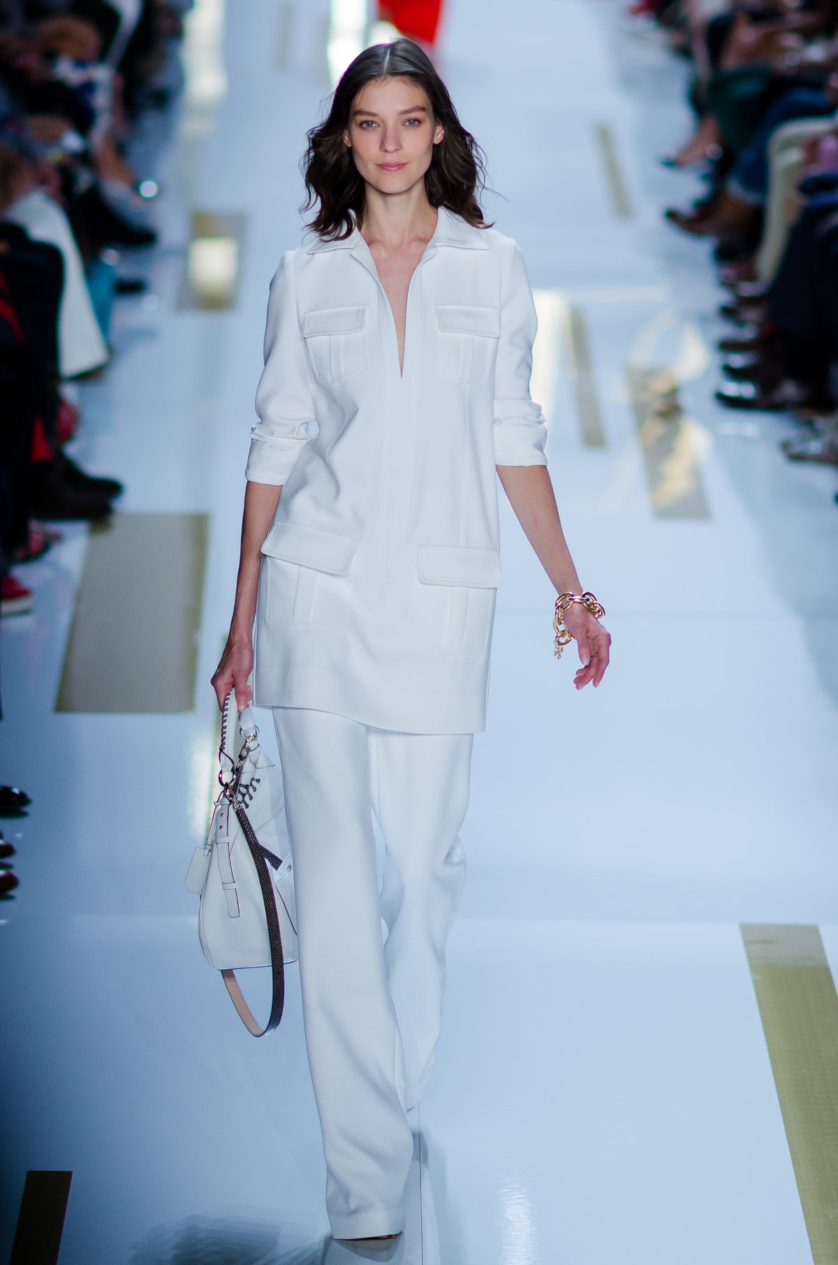Runway model in head-to-toe white: a loose pocketed tunic over wide-leg white trousers, carrying a white handbag