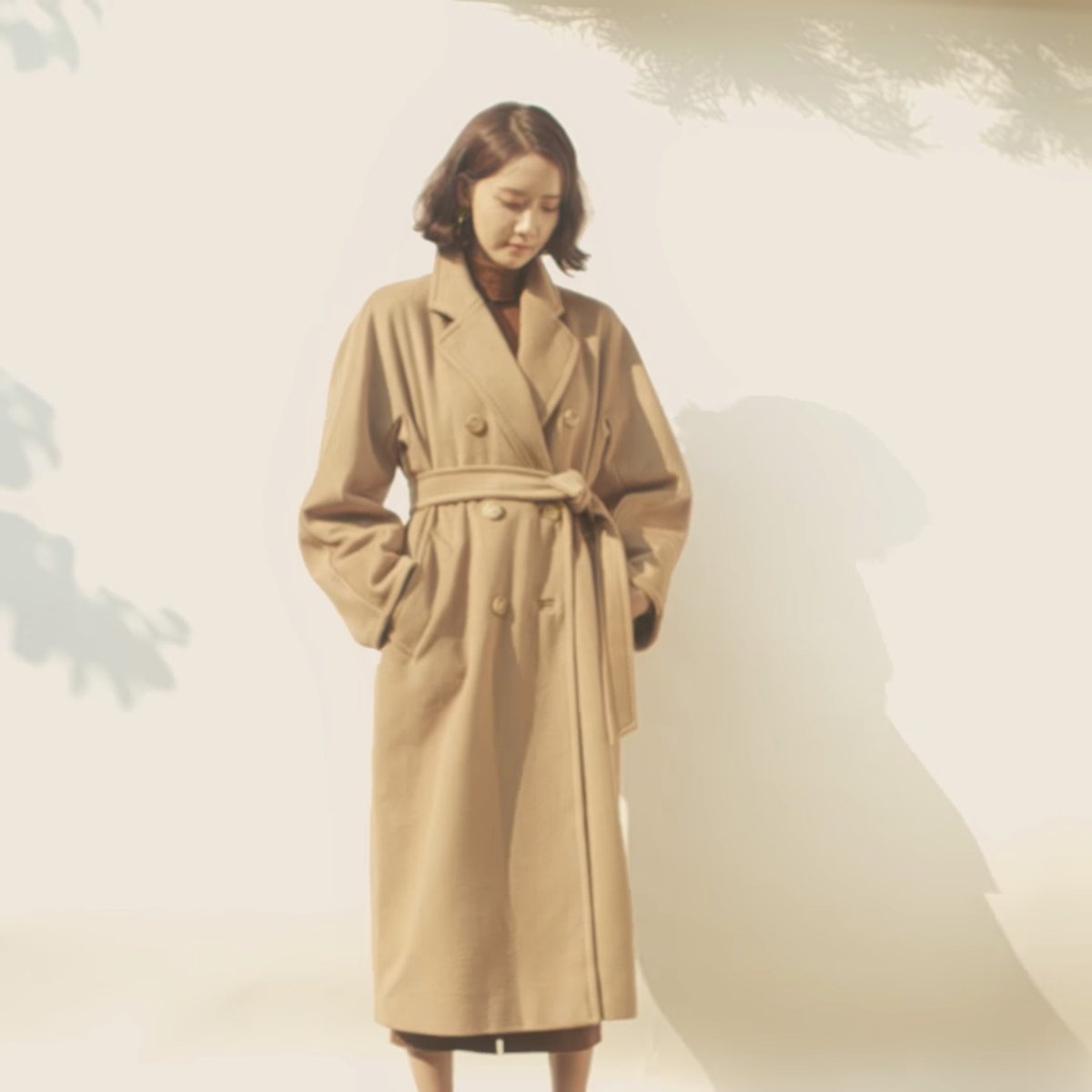 Woman with shoulder-length dark hair wearing a double-breasted camel-tan belted wool coat over a dark brown turtleneck knit, standing against a pale sunlit wall